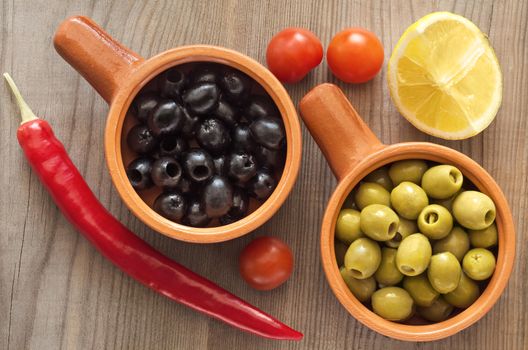 Green and black olives in a ceramic dish. Wooden table, lemon and vegetables.