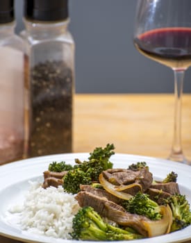 Cooked beef and broccoli stirfry served on rice with a wine glass and spices in the background