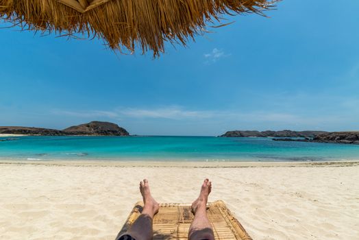 Legs of guy relaxing on sandy beach under umbrella