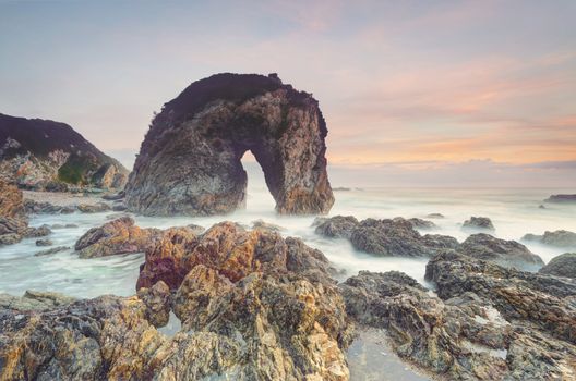 Beautiful coastal seascape with natural rock formations carved out over thousands of years.  Behind the sky lights up in pastel hues of red orange and blue and in the foreground sharp volcanic rocks and rockpools wet with the surging seas