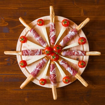 Cutting board with ham rolled in bread-sticks cherry tomatoes and cheese seen from above
