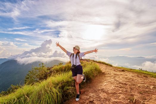 Happy hiker cute teen girl with dslr camera cap eyeglass and backpack stand smiling open arms on peak mountain and sky during sunset background at Phu Chi Fa Forest Park, Chiang Rai, Thailand