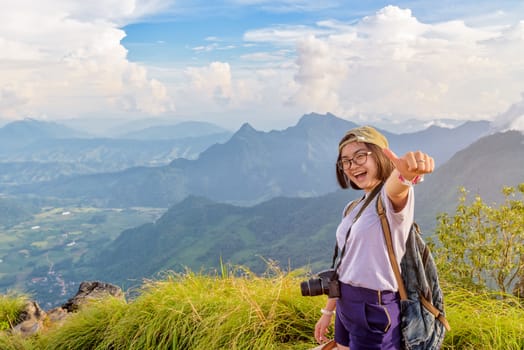 Happy hiker asian cute teen girl with dslr camera cap eyeglass and backpack stand smiling poses thumb up on top of mountain at viewpoint Phu Chi Fa Forest Park in winter, Chiang Rai, Thailand