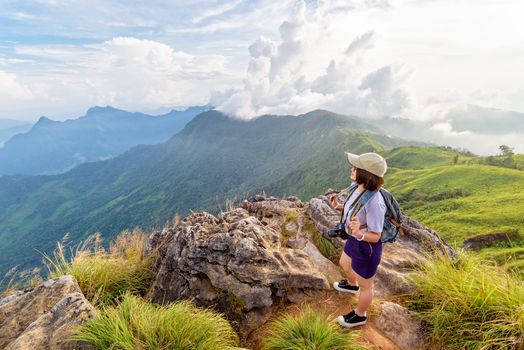 Hiker asian teen girl happy with camera backpack cap and eyeglass looking beautiful landscape nature of mountain range and colourful sky at sunset on Phu Chi Fa Forest Park, Chiang Rai, Thailand
