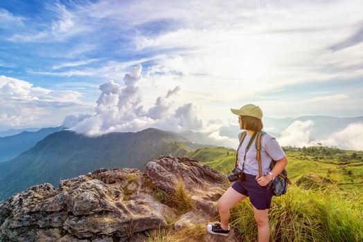 Hiker asian teen girl happy with camera backpack cap and eyeglass looking beautiful landscape nature of mountain range and colourful sky at sunset on Phu Chi Fa Forest Park, Chiang Rai, Thailand