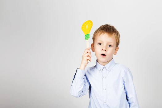 Cute boy with yellow paper lightbulb against a white background. little student holding toy bulb. Cheerful smiling Kid with funny photo props. School concept. Back to School