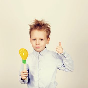 Cute boy with yellow paper lightbulb against a white background. little student gesturing thumb holding toy bulb. Cheerful smiling Kid with funny photo props. School concept. Back to School