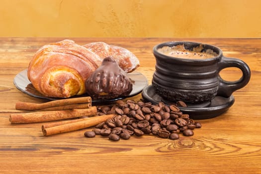 Freshly brewed coffee with milk in the black ceramic cup, scattered beside roasted coffee beans and cinnamon sticks, chocolate truffle and croissant on the glass saucer on the old wooden surface
