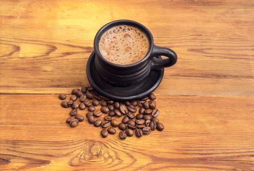 Freshly brewed coffee with milk in the black ceramic cup with saucer and scattered near coffee beans on a surface of an old wooden planks
