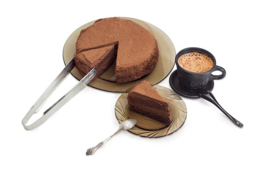 Partly sliced round chocolate cake sprinkled with cocoa powder on a glass dish and kitchen tongs, piece of the cake with spoon on the glass saucer and coffee with milk in black ceramic cup on a light background
