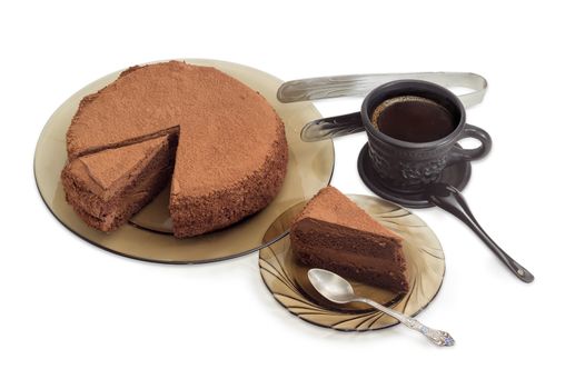 Partly sliced round chocolate cake sprinkled with cocoa powder on a glass dish, piece of the cake with spoon on the glass saucer and black coffee in black ceramic cup on a light background
