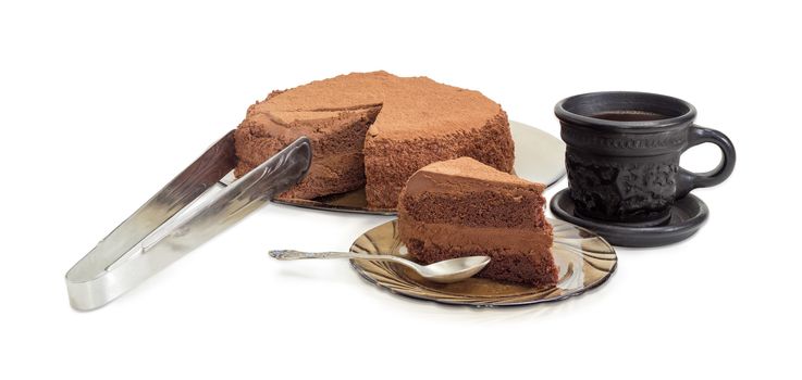 Partly sliced round chocolate cake sprinkled with cocoa powder on a glass dish and kitchen tongs, piece of the cake with spoon on the glass saucer and coffee in the black ceramic cup on a light background
