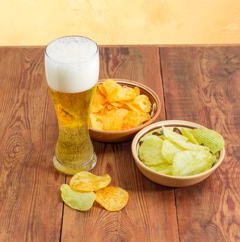 Beer glassware with lager beer, potato chip flavored paprika and wasabi in two different ceramic bowls and several potato chips separately beside on an old wooden surface
