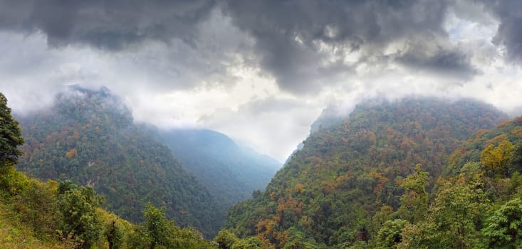 Panorama of the mountain gorge with the slopes covered with autumn forest and storm clouds above him in the Himalayas
