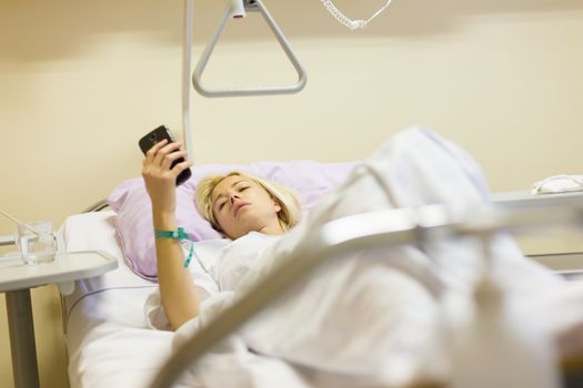 Bedridden female patient lying in hospital bed, using mobile phone while recovering after surgery.