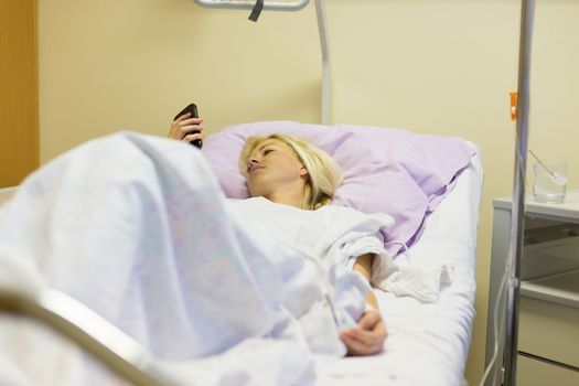 Bedridden female patient lying in hospital bed, using mobile phone while recovering after surgery.