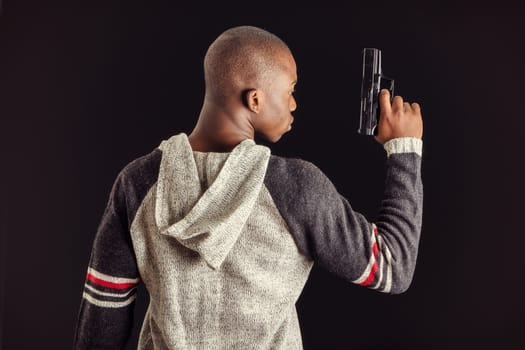 Young handsome black man holding a hand gun, on dark background in studio, seen from the back