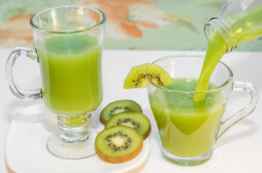 Fresh kiwi juice is poured from the bottle into the Cup, which stands on the table, pieces of fruit nearby. Selective focus.