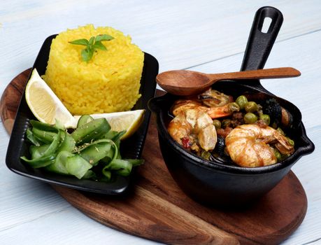 Arrangement of Delicious Seafood Curry with Saffron Rice and Cucumber Salad on Serving Board with Wooden Spoon closeup on Wooden background