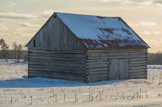 Pioneer log cabin barn in Eastern ontario in winter landscape.  Sun is setting, clouds in the sky ansd corn stalks stick up in the snowy field.