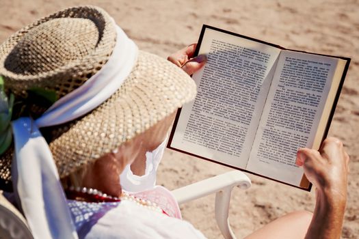 adult woman in a hat on a beach reading a printed book.