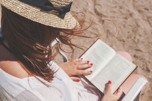 Young adult woman with a hat on the beach reading a book.