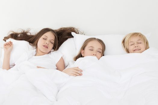 Three beautiful children sleeping in bed under one white blanket