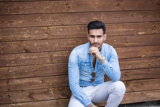 Attractive young man sitting against wood wall, looking at camera