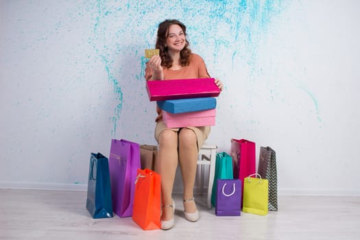 Happy smiling woman after shopping sits with colourful paper bags, boxes showing banking credit card