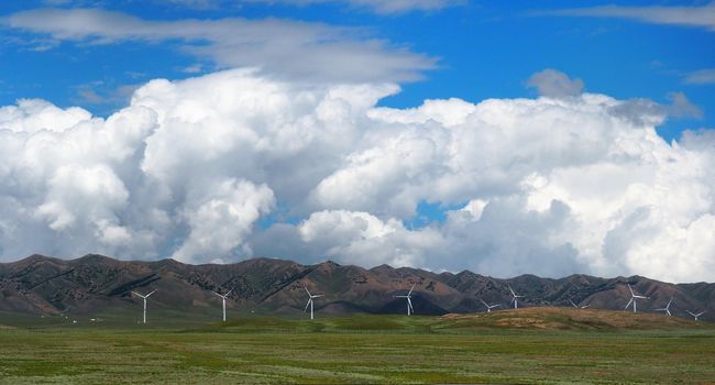 Landscape Of Wind turbines Or Windmills On Green Grass Fields With Mountain Ranges And Cloudy Blue Sky Background, Alternative Energy In Xinjiang, China