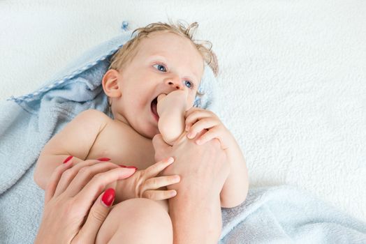 Cute happy beautiful infant boy playing with his foot with wet hair sitting in hothouse bath light blue fluffy towel white background, horizontal photo.