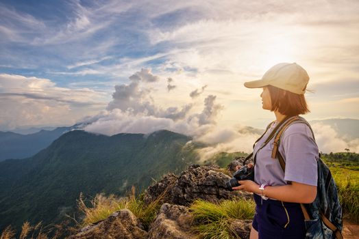 Hiker asian young woman happy with camera backpack and caps looking beautiful landscape nature of mountain and colourful sky at sunset on viewpoint Phu Chi Fa Forest Park, Chiang Rai, Thailand