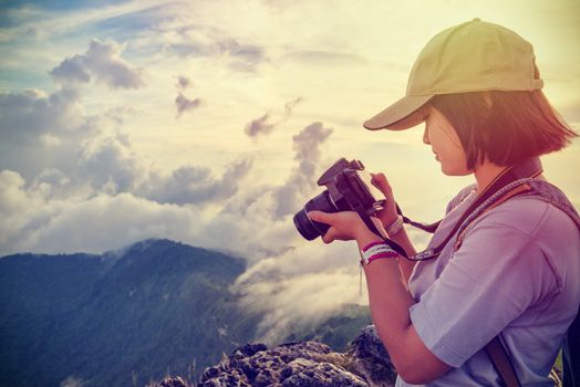 Vintage style hiker teens girl wear caps looking photo at digital camera is beautiful landscape natural of sierra and sky during sunset on mountain in Phu Chi Fa Forest Park, Chiang Rai, Thailand