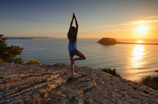 A woman standing on a rock looking out to the sea at sunrise doing tree pose variation, yoga, meditation, Vrksasana