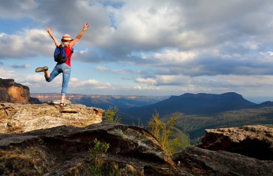 Happy female hiker, traveller, feeling, elation, joy, success, or other positive mood or feeling of wellbeing.  She is high on a rocky outcrop overlooking the Jamison Valley in the Blue Mountains of Australia with Mount Solitary in shade and far in the distance are King Tablelands.