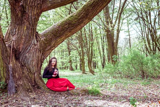 mystical witch woman in red dress sitting under a old tree in the wood