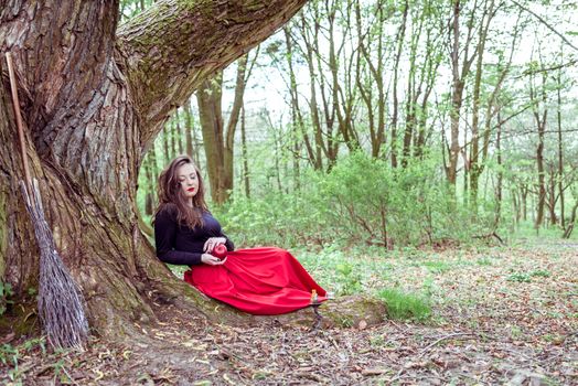 mystical witch woman in red dress sitting under a old tree in the wood