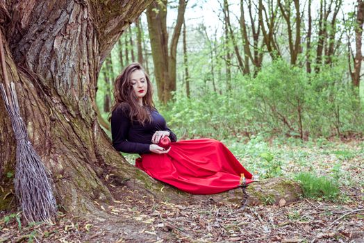 mystical witch woman in red dress sitting under a tree and holding an apple in the park