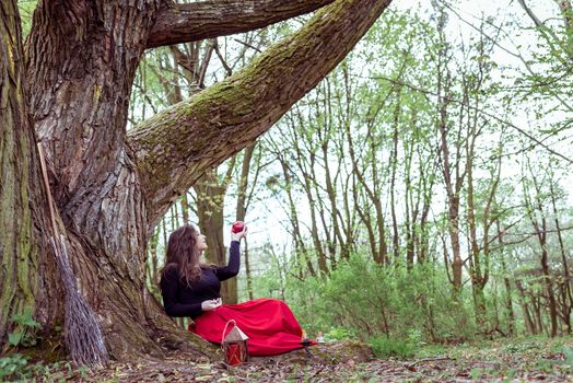 mystical witch woman in red dress sitting under a old tree in the wood