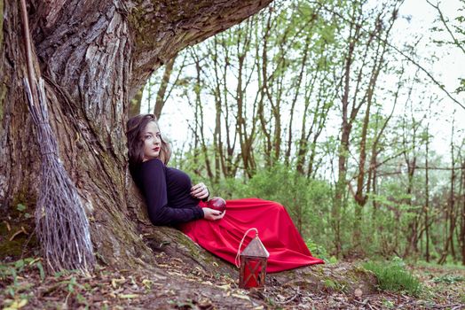 mystical witch woman in red dress sitting under a old tree in the spring park