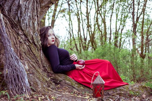 mystical witch woman in red dress sitting under a old tree in the spring park