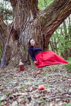 mystical witch woman in red dress sitting under a old tree in the spring park