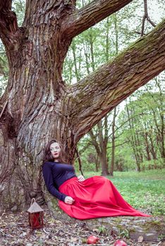 mystical witch woman in red dress sitting under a old tree in the wood