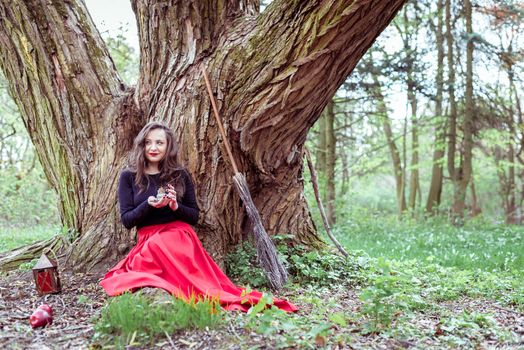 mystical witch woman in red dress sitting under a old tree and holding a candle in a spring forest