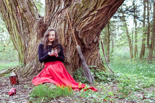 mystical witch woman in red dress sitting under a old tree and holding a candle in a spring forest
