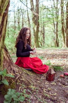 mystical witch woman in red dress sitting under a old tree and holding a candle in a spring forest