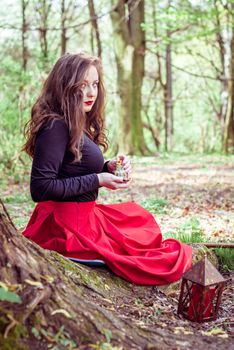 mystical witch woman in red dress sitting under a old tree and holding a candle in a spring forest