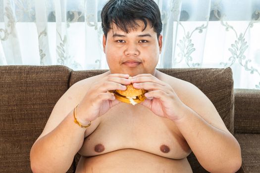 Asian fat man eating hamburger seated on armchair