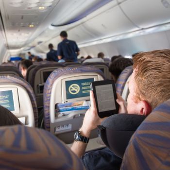 Rear view of airplane cabin with unrecognizable male passenger reading e-book on electronic reader during flight.