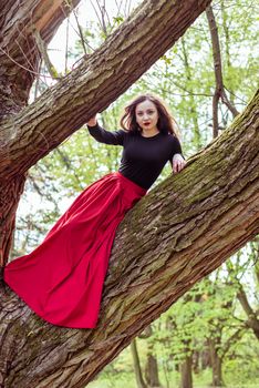 beautiful woman in a red dress sitting on a trunk in the spring forest